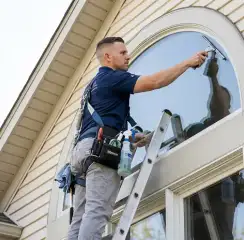 Professional window cleaner on a ladder using a squeegee to clean an exterior arched window, New Haven, CT.